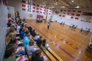 Engaged students attending a school assembly in a gymnasium with banners and American flags.