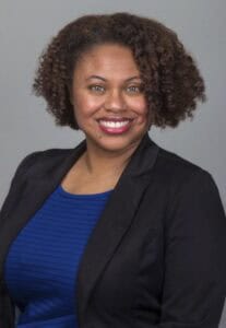 A woman with curly hair smiling in professional attire against a neutral background.