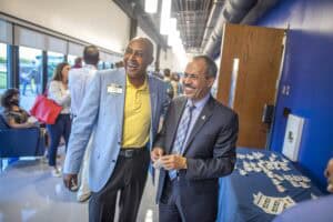 A smiling man in a yellow shirt and blue blazer talking with another man in a suit at a professional.