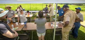Community meeting outdoors under a tent with diverse participants discussing agricultural topics.