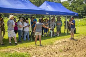 Community outdoor event with people gathered under blue canopy tent on sunny day.