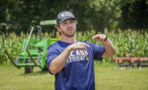Young man in a blue CAES Gaggies t-shirt outdoors with farm equipment in background.
