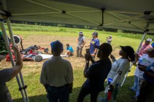 Community gathering at outdoor event with people under a canopy and a tractor in the background.