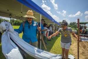 Planting volunteers working together outdoors at a community event, under a blue canopy on a sunny d.