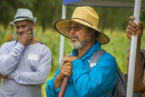 Elderly man with straw hat and blue shirt holding a walking stick outdoors.