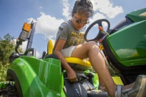 Green tractor with a young woman sitting on the seat, adjusting controls outdoors under a partly clo.