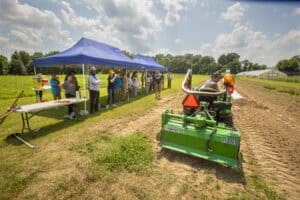 Agricultural equipment demonstration at outdoor event with attendees and a green tractor attachment.