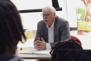 Senior man speaking during a meeting or interview at a desk in an office setting.