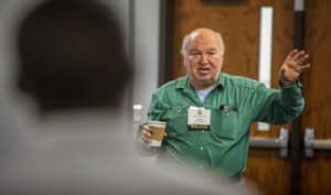 A man speaking at a conference or seminar, holding a coffee cup and wearing a name badge.