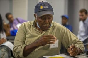 Man wearing a cap and beige shirt, focused on writing at a conference table.