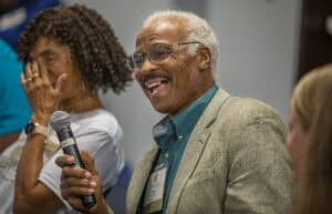 A smiling senior man speaking into a microphone at a conference or event.