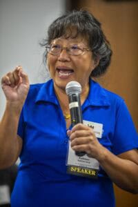 Empowered woman speaker at conference, wearing blue shirt with "SPEAKER" badge, passionately address.