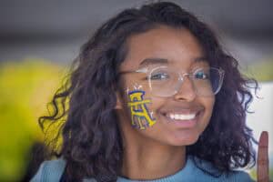 Smiling young woman with glasses and CAES logo sticker on her face at CAES News event.