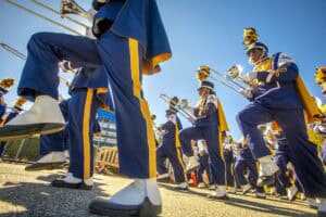 Marching band performers in uniform during outdoor parade, playing instruments under clear blue sky.