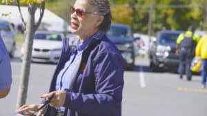A woman in sunglasses and a blue jacket standing outdoors in a parking lot.