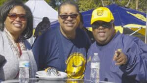 Group of three smiling people at an outdoor event with yellow and blue tents.
