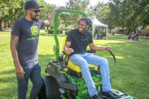John Deere lawn tractor with two men outdoors in a park setting, one sitting on the tractor and the.