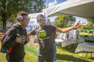 Engaging outdoor event with two men discussing under a tent, promoting John Deere equipment.