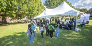 Community outdoor event with a white tent, people gathering and socializing in a park setting.