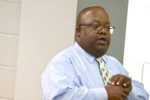 Professional man speaking at a conference or seminar, wearing glasses, a light blue shirt, and a yel.