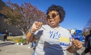 Bright smiling woman with curly hair wearing sunglasses and a gray T-shirt outdoors during daytime.