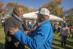 Smiling men greeting each other at outdoor community event on sunny autumn day.