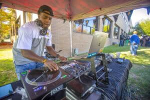 DJ performing at an outdoor event with a laptop and sound equipment under a tent.