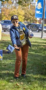 A woman wearing a University of Michigan jacket and cap, holding a donation box outdoors during fall.