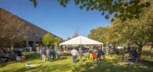 Outdoor community event with people gathering under a white tent on a sunny day, surrounded by trees.
