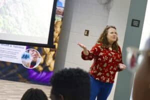 Healthy woman giving a presentation in a classroom or seminar setting with a large screen behind her.