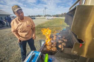 A person grills burgers as flames rise from the grill, surrounded by a scenic outdoor setting.