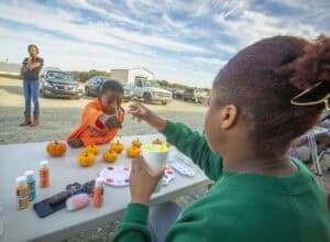 A child paints mini pumpkins while an adult hands them a paintbrush at an outdoor table.