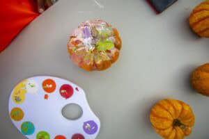 A painted pumpkin sits on a table beside a palette of colorful paints and several small pumpkins.
