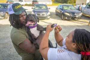 A person holds a happy black dog while another paints its toenails in a parking lot.