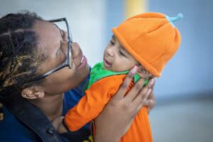 A smiling woman holds a baby dressed in an orange pumpkin outfit with a matching hat, both looking joyful.