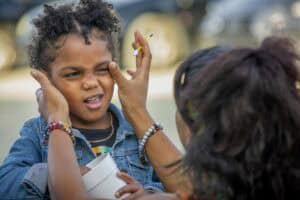 Child squints playfully as an adult applies makeup, surrounded by sunny outdoor scenery.
