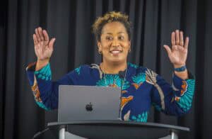 Empowered woman speaking at a conference with a laptop, raising hands in a confident gesture.
