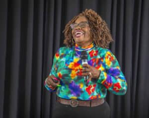 Laughing woman with curly hair and glasses speaking into a microphone at a conference or event.
