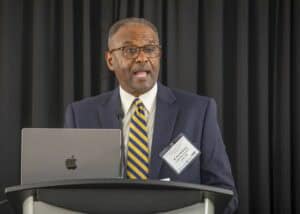 African American man speaking at conference with laptop and microphone.