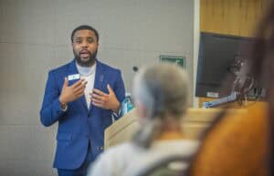 A man in a blue suit giving a presentation in a conference room.
