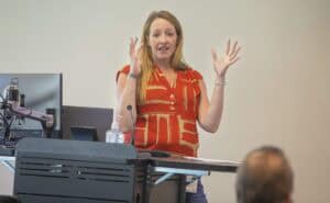 A woman speaking at a conference or seminar, gesturing with her hands.
