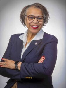 A woman in professional attire smiling with crossed arms, wearing glasses and a smartwatch.