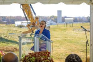 A woman speaking at an outdoor event with a construction excavator in the background.