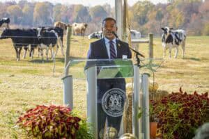 Farmers' conference with a speaker at a podium outdoors, surrounded by cows and autumn foliage.