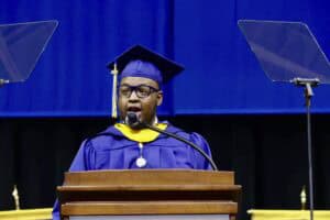 Graduation speaker at ceremony in cap and gown delivering speech at podium.