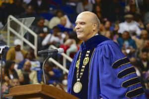 Graduation ceremony with a smiling man in academic regalia speaking at a podium.