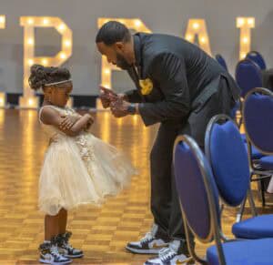 A man in a suit talks to a girl in a dress and sneakers, both looking serious in a decorated venue.