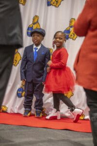Two children pose on a red carpet: a boy in a suit and a girl in a red dress with stylish sneakers.