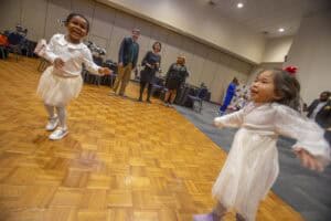 Two young girls in white dresses joyfully dance on a wooden floor, with adults enjoying the event in the background.