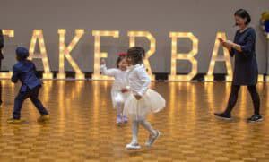 Children dance joyfully in a bright hall with illuminated letters spelling "SPEAKER BALL." An adult smiles nearby.
